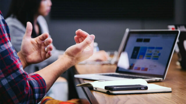 Close-up of hands expressing a point while a laptop and smartphone rest on a notebook during a meeting