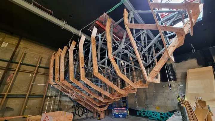 Wooden and metal framework of a large climbing wall suspended inside an unfinished industrial space