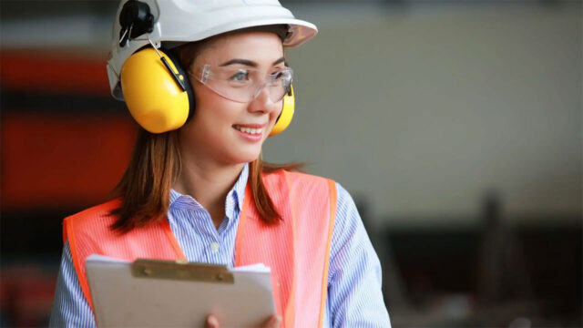 Person in safety vest, hard hat, ear protection, and glasses holding clipboard