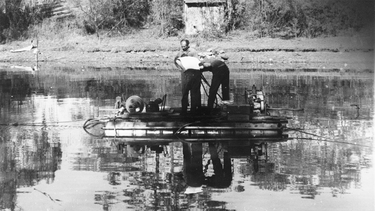 Two men working on a small floating platform with equipment on a calm body of water