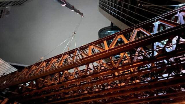 Crane lifting large steel beams at night in an urban construction site with tall buildings