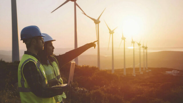 Two workers in safety vests and helmets inspecting wind turbines at sunset, one pointing ahead