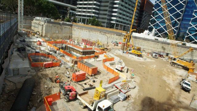 Urban construction site with orange barriers, cranes, trucks, and workers near modern office buildings