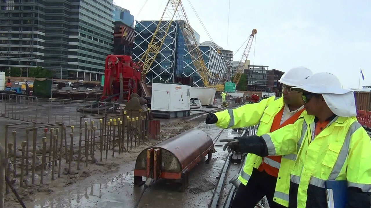 Two construction workers in yellow jackets and white helmets discussing at muddy urban construction site