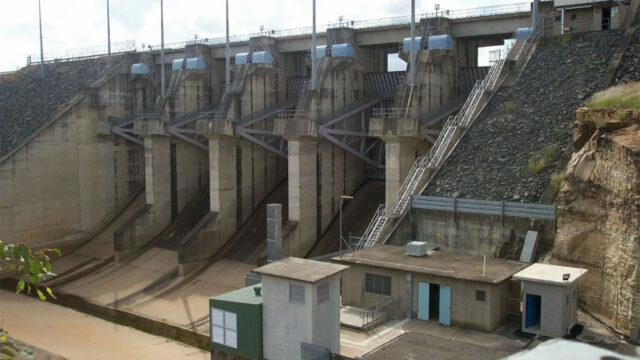 Concrete dam with spillways, control gates, and adjacent buildings by rocky hillside