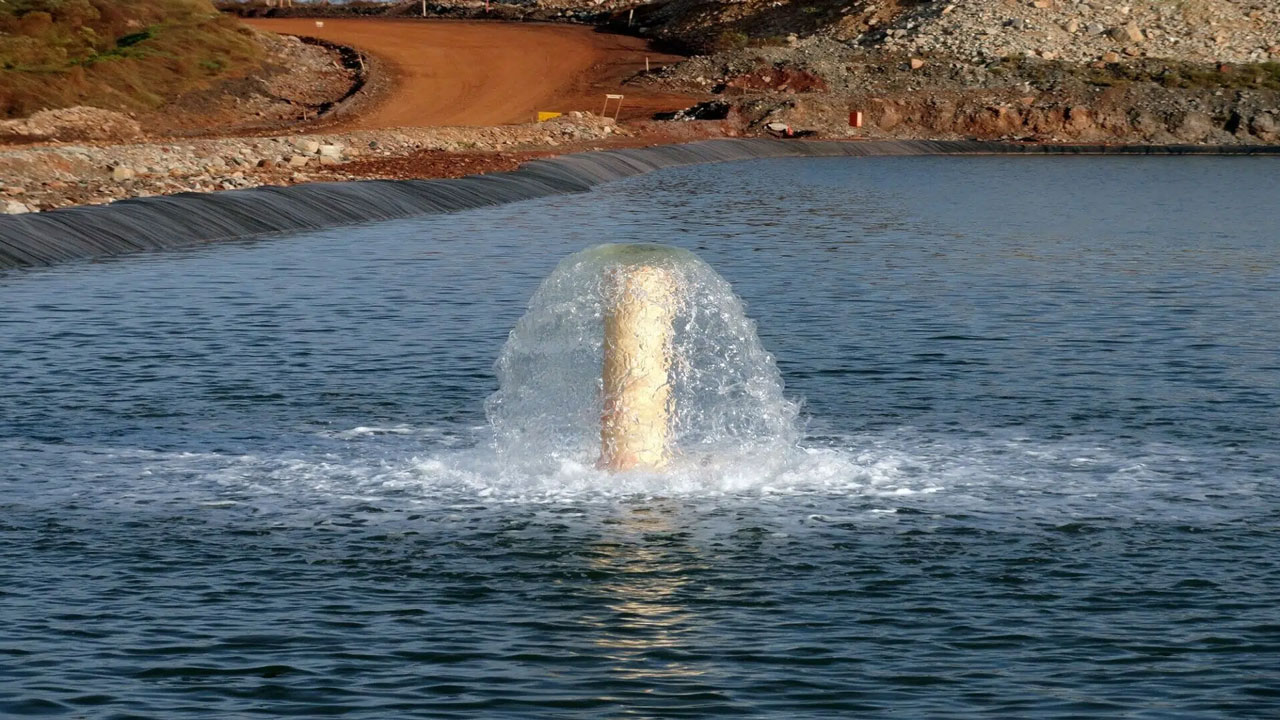 Water flowing from a pipe into pond near a dirt road and rocky embankment