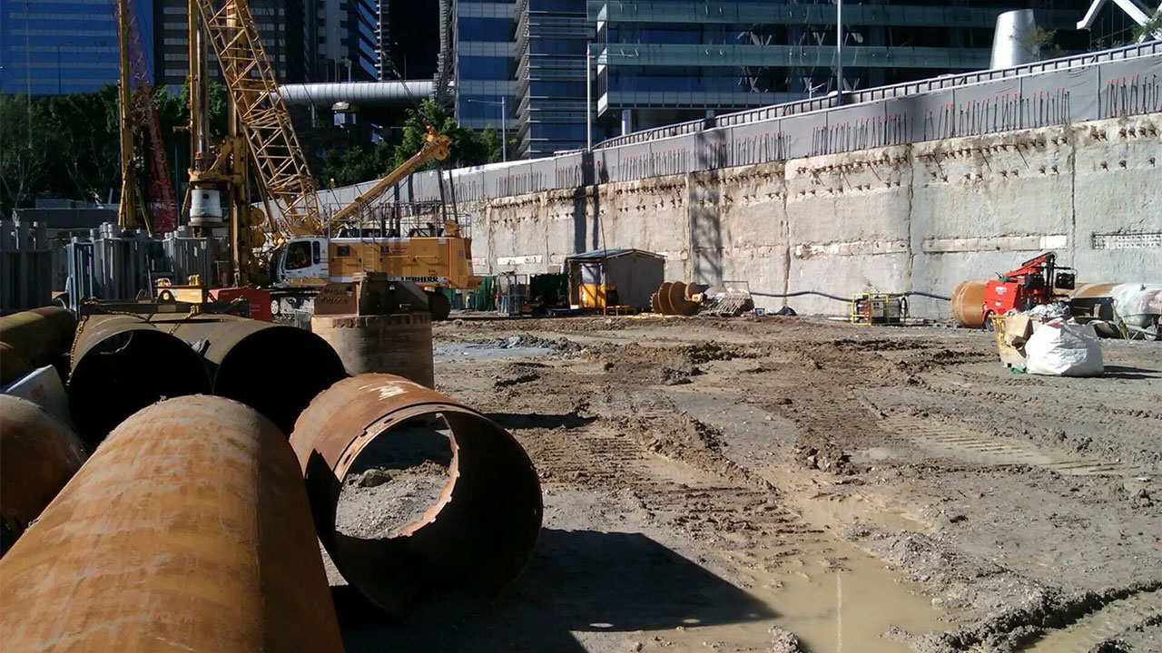 Large construction site with heavy machinery, rusty pipes, and muddy ground near concrete retaining wall