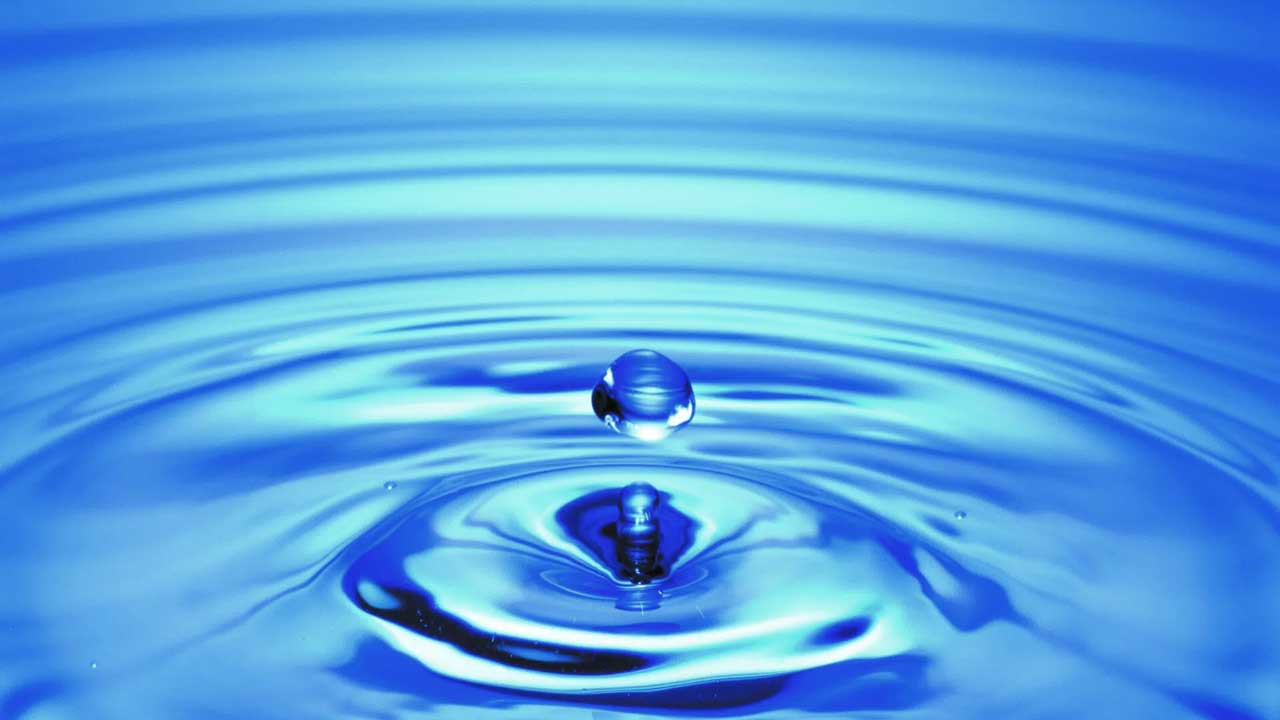 A close-up of a single water droplet above the surface creating ripples in blue water