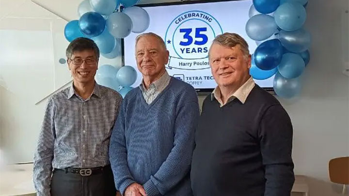 Harry Poulos standing between two colleagues in front of a banner with text that reads "Celebrating 35 years"