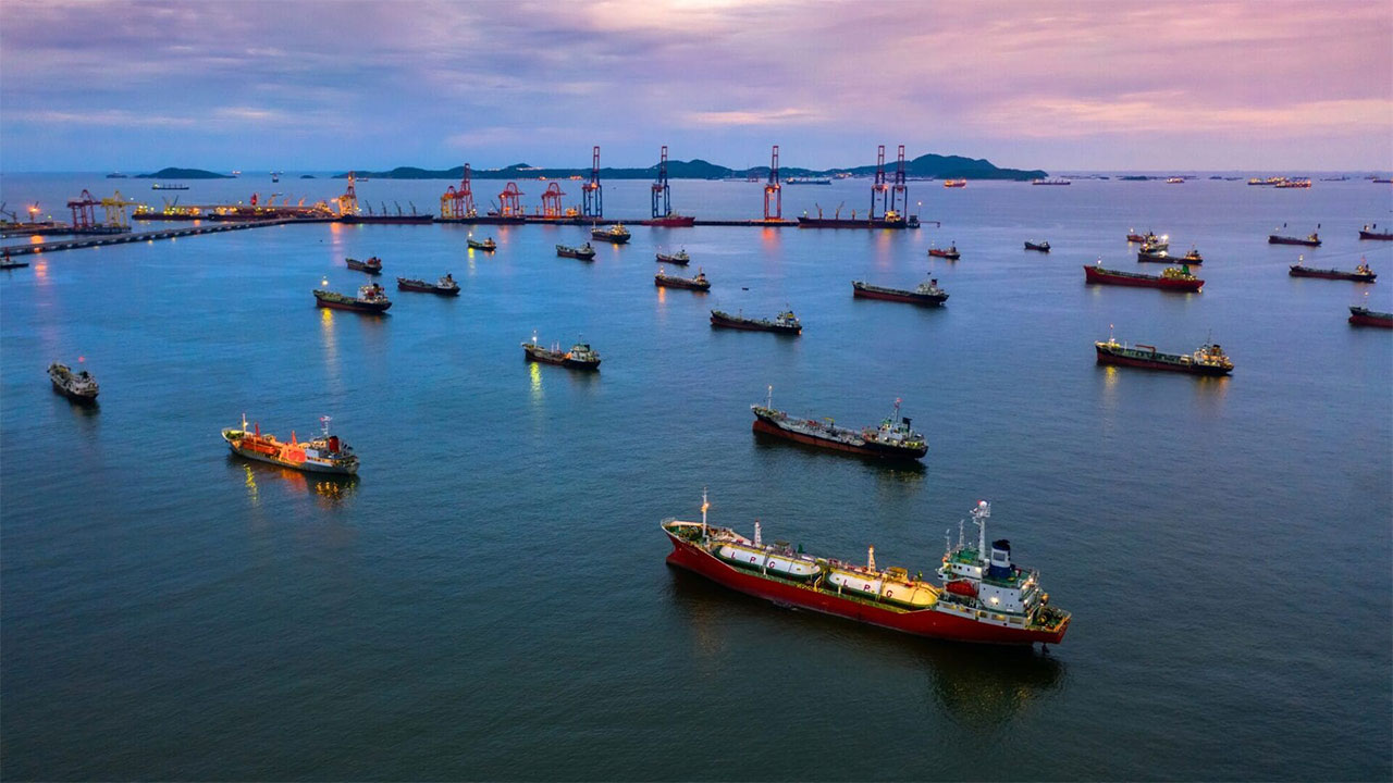 Cargo ships anchored near a busy port at dusk with cranes and distant islands