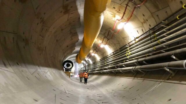 Interior view of the Melbourne Metro Tunnel under construction with workers walking inside the large concrete tunnel next to pipes and ventilation ducts