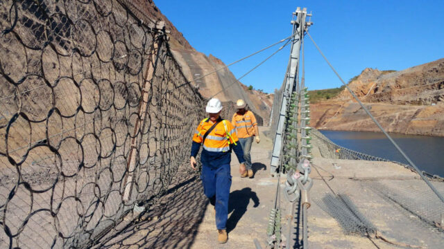 Two workers in safety gear walking on a dam structure near the rocky hillside and water