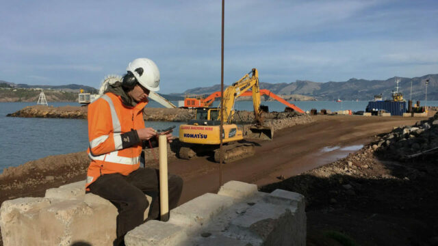 Construction worker in orange safety gear and white helmet using a device while sitting near heavy machinery and water with mountains in the background