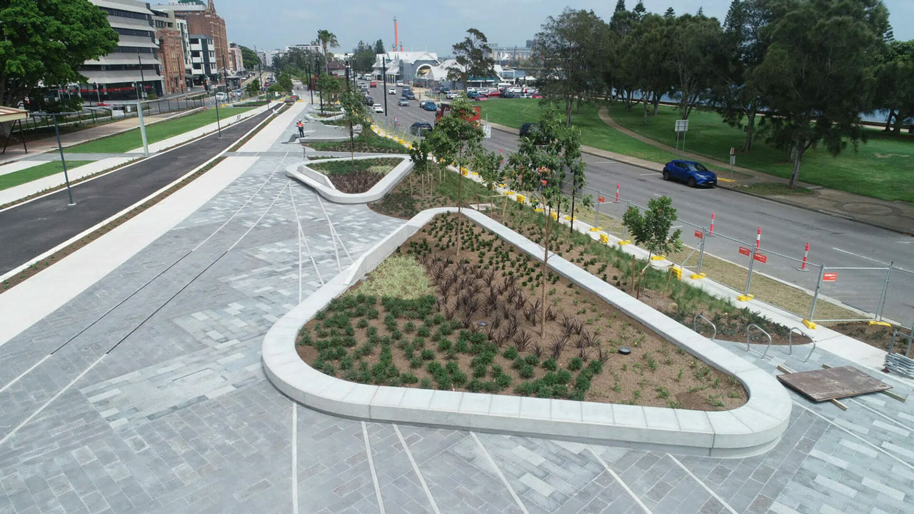 Urban landscaped area with triangular garden beds, paved walkways, road, and parked cars nearby