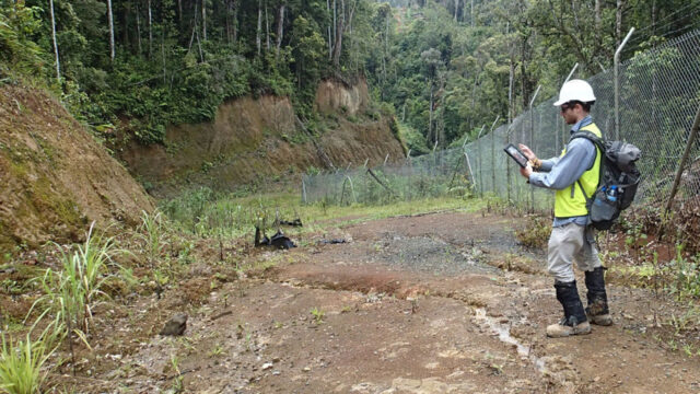Worker in safety gear using a tablet to survey the forested terrain within a fenced area