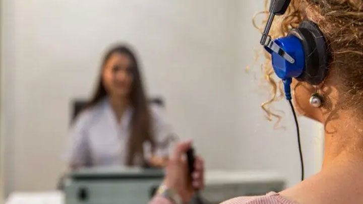 Person wearing blue headphones facing another person in a white shirt in a blurred background holding a single button controller