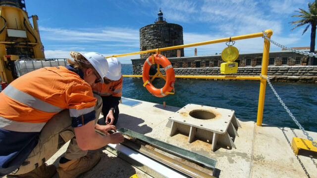 Two workers in safety gear inspecting equipment on a dock by water and stone building in the background