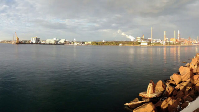 Industrial waterfront with factories, smokestacks, and cranes across calm water, and a rocky shoreline in the foreground