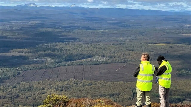 Two people in yellow reflective vests stand on a mountaintop, overlooking a vast forested valley with distant mountains