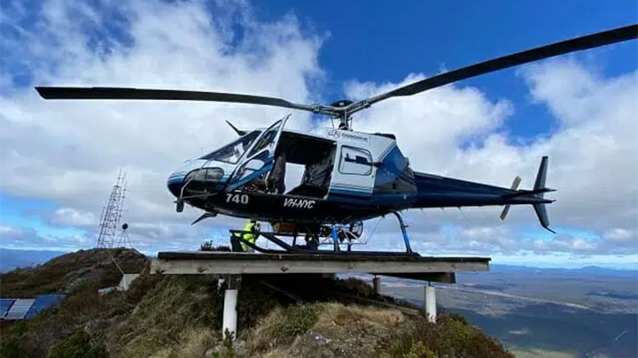 A blue-and-white helicopter (VH-NYC) on a wooden landing platform atop a hill, worker in yellow vest and small radio tower