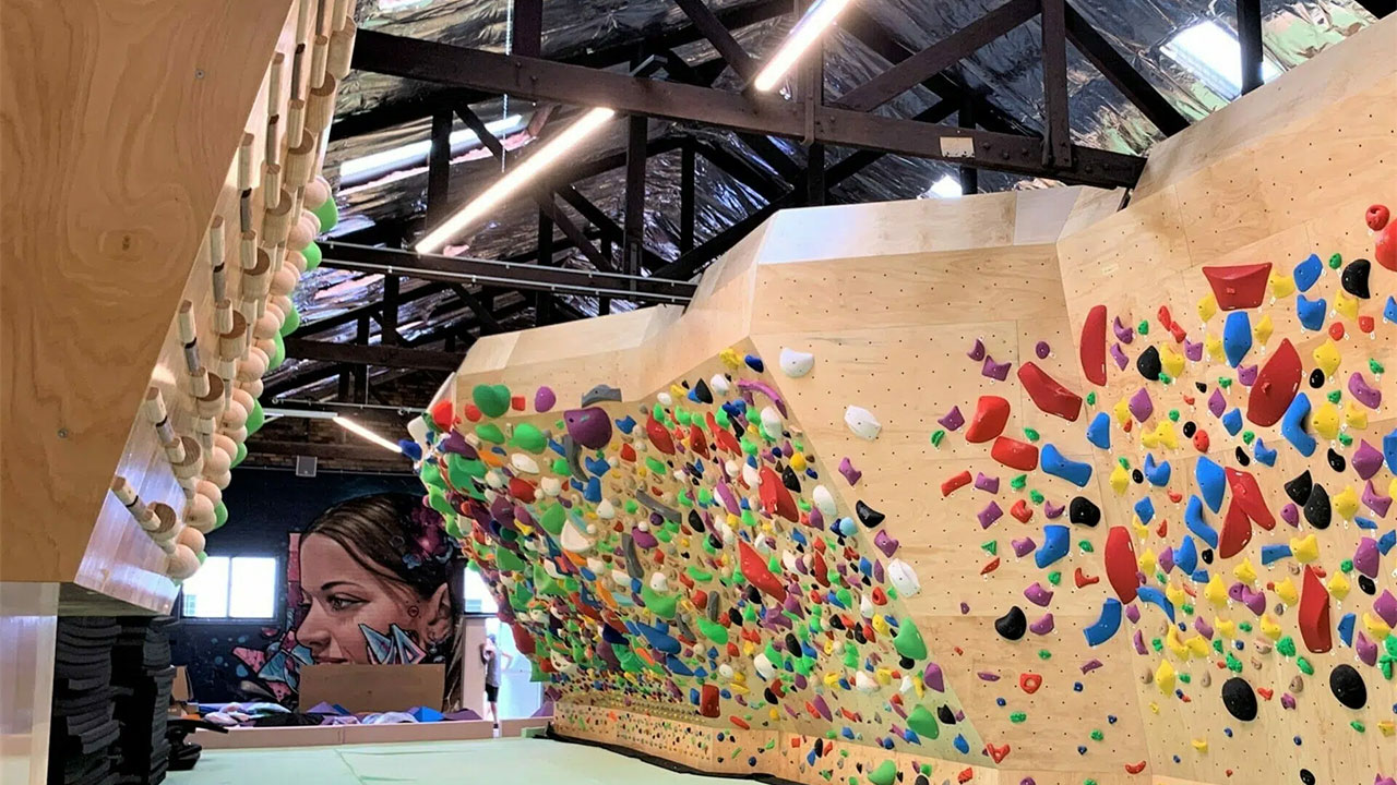 Indoor bouldering wall with multicolored holds, campus board, muraled face, under construction beneath exposed ceiling beams