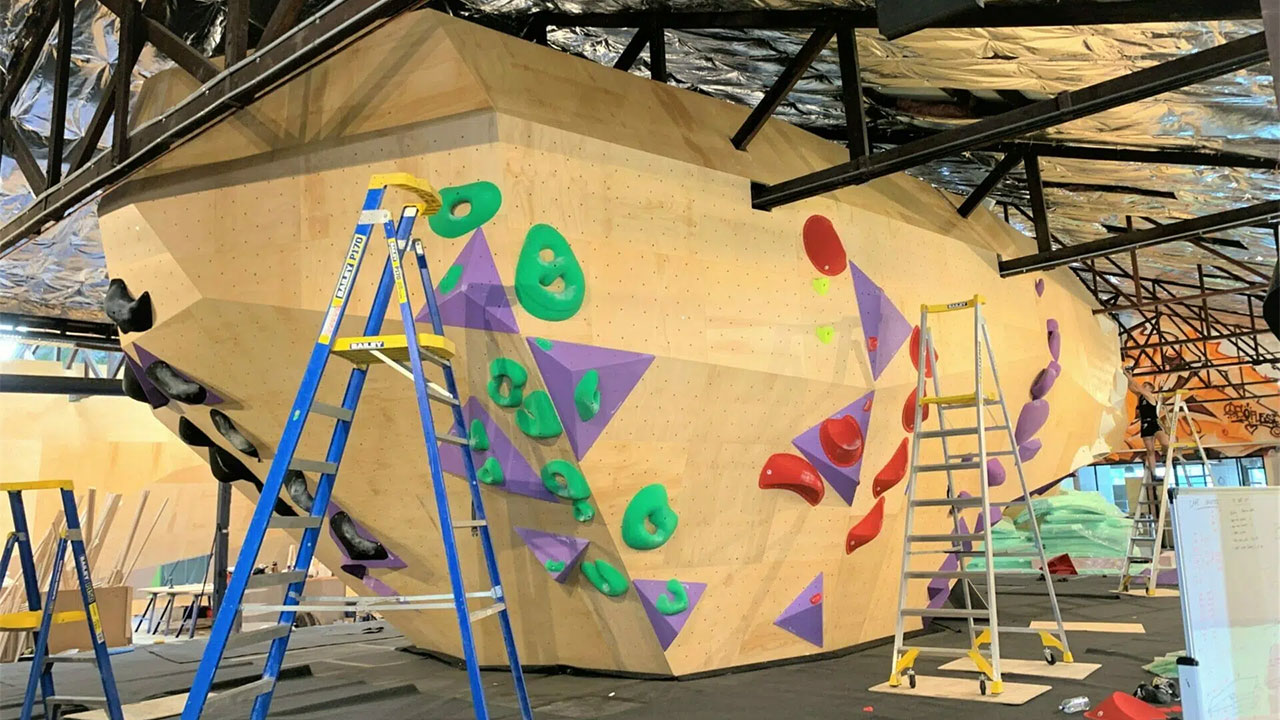 Angular climbing wall with green, red, purple holds, ladders and mats during installation in gym